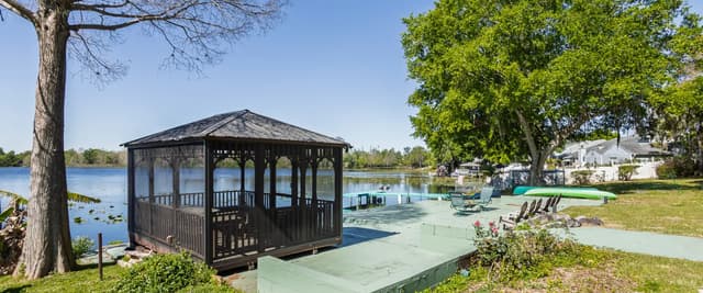 Waterfront property with black gazebo, dock, and lawn chairs overlooking calm lake with trees and houses